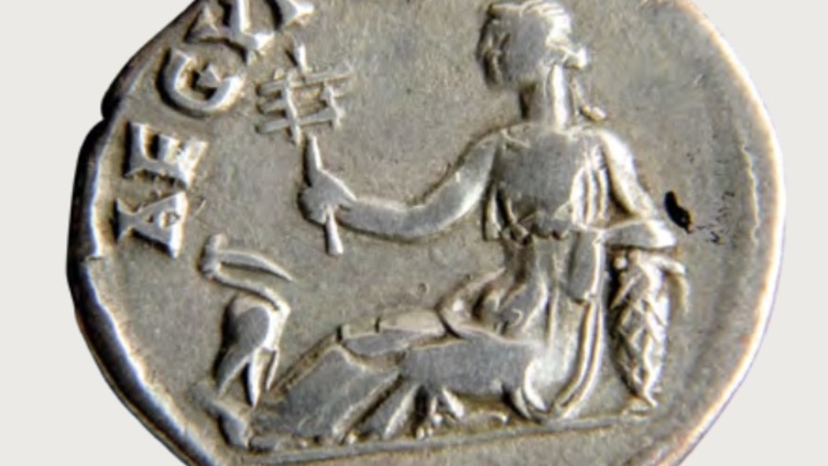 An ancient silver coin depicts a seated figure holding a scepter. The figure is draped in robes and sits with one leg extended.